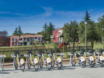 Estación de alquiler de bicicletas con varias bicicletas frente a un parque y edificios al fondo.