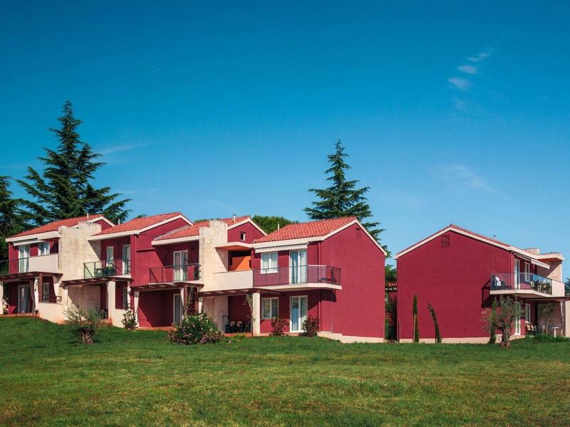 Case vacanza rosse con balconi e prato verde sotto un cielo azzurro.