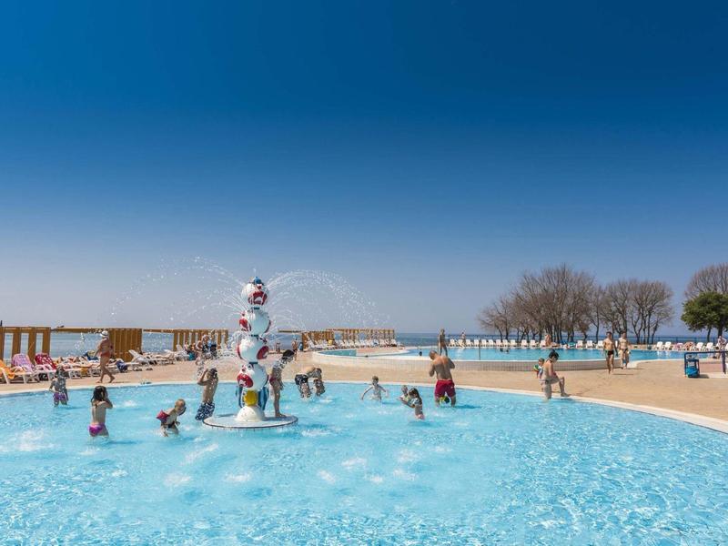 Bambini che giocano in una piscina esterna di un hotel sotto un sole splendente e un cielo azzurro.
