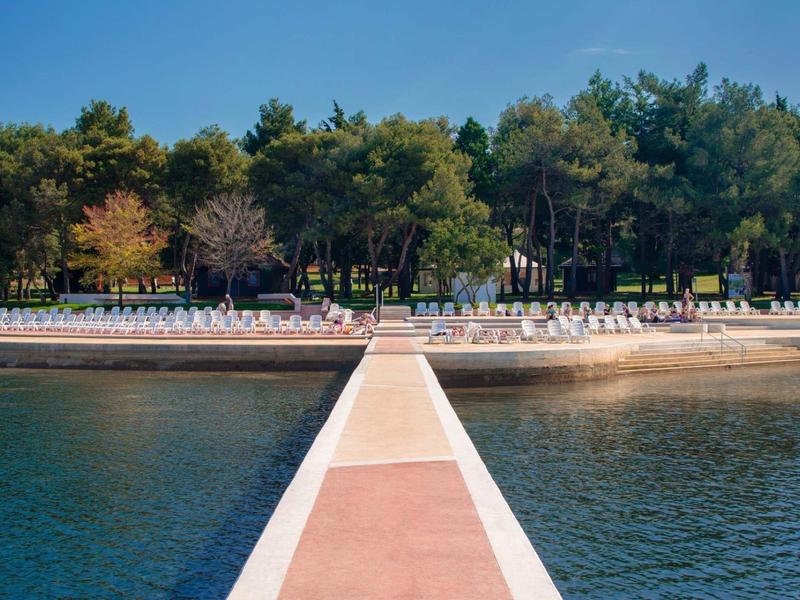 Un pontile conduce a una spiaggia sabbiosa con sdraio e alberi sullo sfondo.
