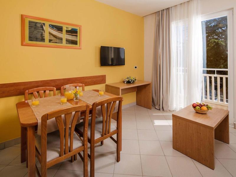 Sala da pranzo luminosa con tavoli e sedie in legno, pareti gialle e balcone con vista sugli alberi.