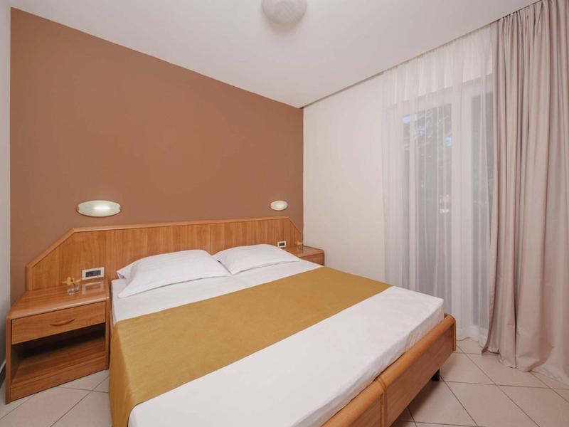 Camera d'hotel luminosa con letto matrimoniale, comodino e grandi tende alla finestra.
