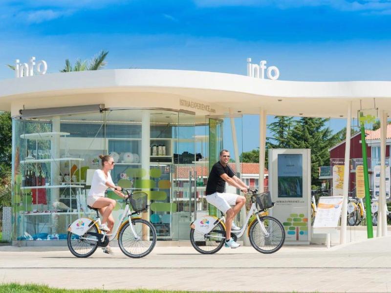 Due persone pedalano accanto a un moderno chiosco informativo con vetrata.