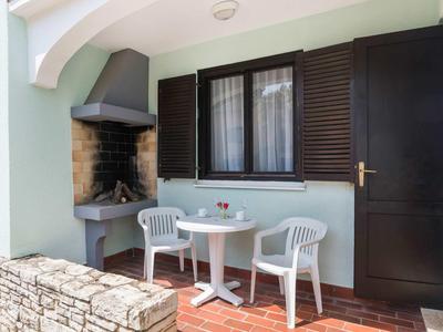 Pequeña zona exterior con dos sillas blancas, mesa redonda y barbacoa junto a una ventana con persianas marrones.