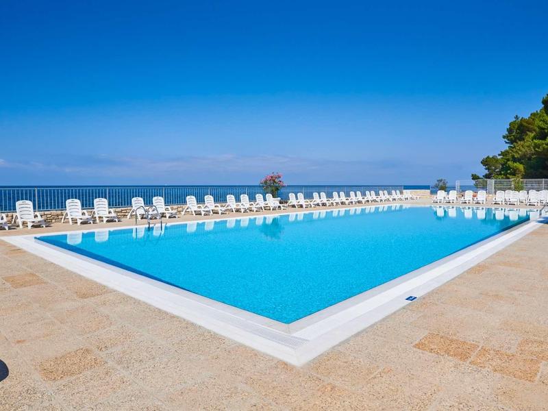 Grande piscina rettangolare con sedie a sdraio bianche e cielo blu limpido vicino al mare.