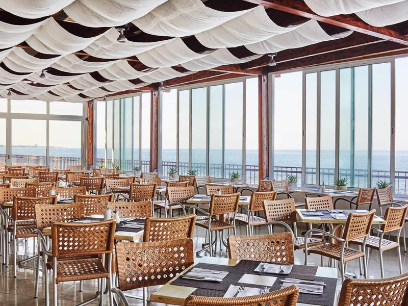 Ristorante luminoso con sedie in vimini e grandi finestre con vista sul mare.