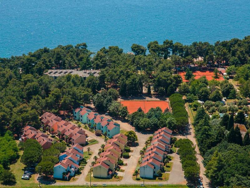 Vista aerea di un resort con case a schiera, alberi verdi e campi da tennis vicino al mare.