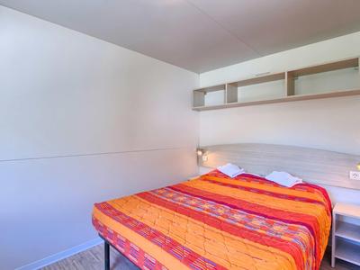 Een eenvoudige slaapkamer met een tweepersoonsbed, oranje-rode beddengoed en een wandplank.