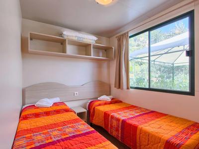 Kleine slaapkamer met twee eenpersoonsbedden, oranje-rode dekens en een raam met uitzicht op groen.