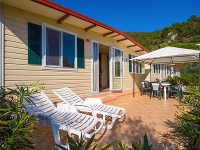 Haus mit gelber Holzfassade, Terrasse mit weißen Liegestühlen und Sonnenschirm, grüne Pflanzen.