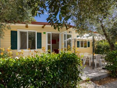 Kleines Ferienhaus mit Terrasse, von Büschen und Bäumen umgeben, sonnig und einladend.
