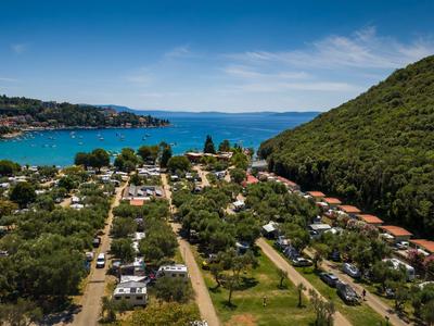 Grüner Campingplatz an der Küste mit Blick auf das blaue Meer und bewaldete Hügel.