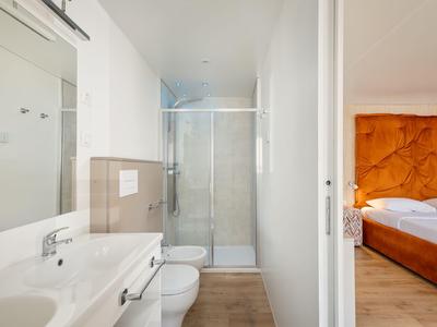 Helles Badezimmer mit zwei Waschbecken, Toilette und Duschkabine neben einem Schlafzimmer.