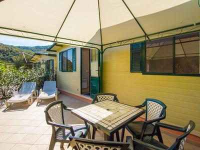Terrasse mit Stühlen, Tisch und Liegestühlen vor einem gelben Haus in sonniger Landschaft.