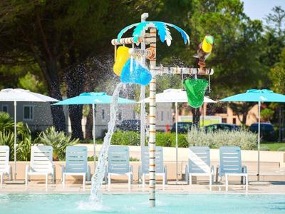 Kolorowy plac zabaw wodny z parasolami i leżakami obok basenu pod drzewami.