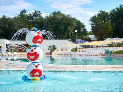 Kolorowa rzeźba klauna z fontannami wodnymi w basenie otoczonym leżakami i zielonymi drzewami.