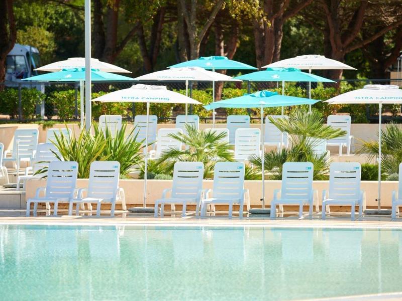 Obszar basenu z białymi leżakami i niebieskimi parasolami pod drzewami.