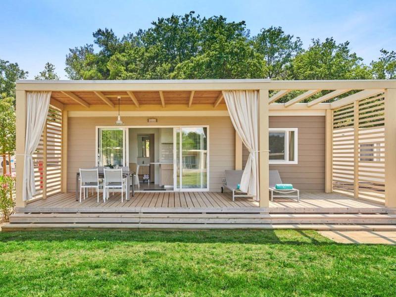 Nowoczesny domek wakacyjny z drewnianym tarasem, miejscem do siedzenia i zasłonami w zielonym otoczeniu.
