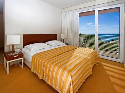 Světle hotelový pokoj s manželskou postelí, nočními stolky a výhledem na moře přes velká okna.