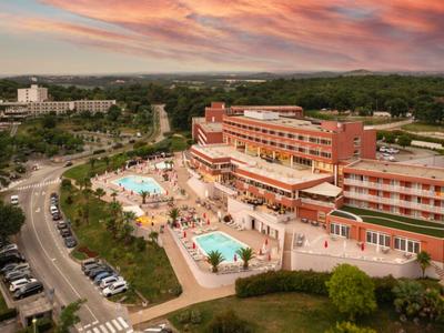 Velký hotelový komplex s bazény, parkovištěm a zalesněným pozadím při západu slunce.