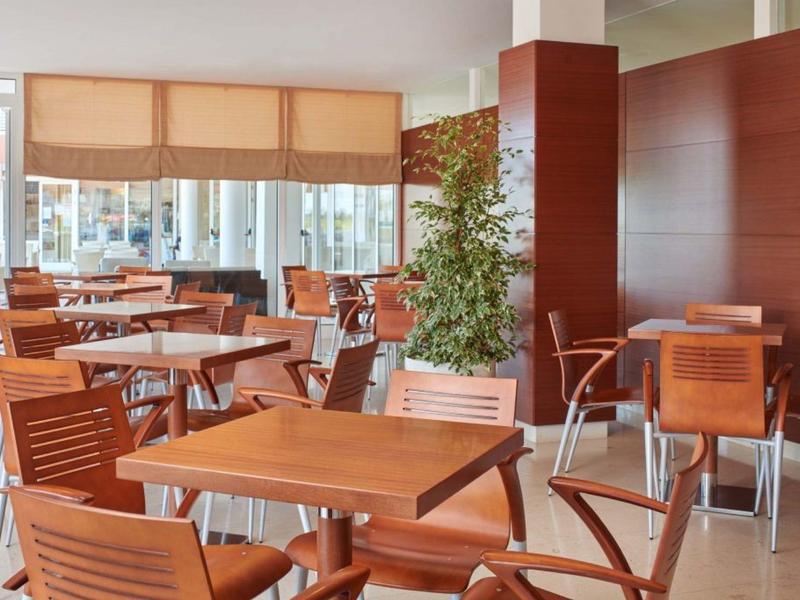 Světlá moderní restaurace s dřevěnými židlemi a stolky, velkými okny a rostlinami.