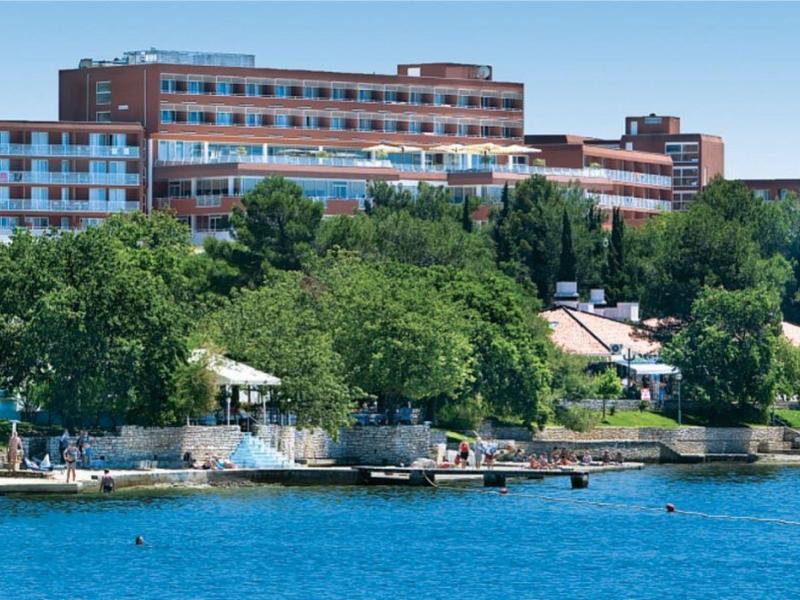 Velká hotelová budova za stromy u břehu s modrou vodou v popředí.