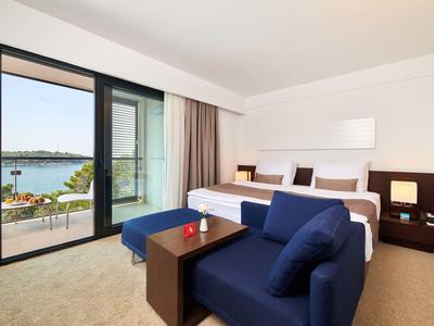 Chambre d'hôtel lumineuse avec lit king-size, canapé bleu et terrasse avec vue sur l'eau.