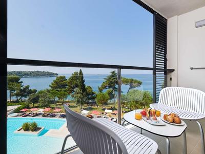 Balcon avec chaises blanches et petite table, vue sur piscine, palmiers et mer sous un ciel clair.