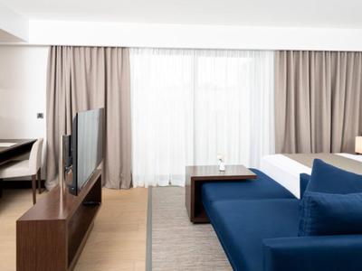 Chambre d'hôtel moderne avec lit, canapé bleu, TV et bureau devant une grande fenêtre avec rideaux.