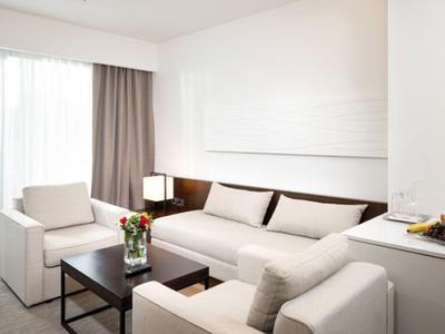 Chambre d'hôtel moderne avec mobilier blanc, canapé, fauteuils et grande fenêtre avec rideaux.