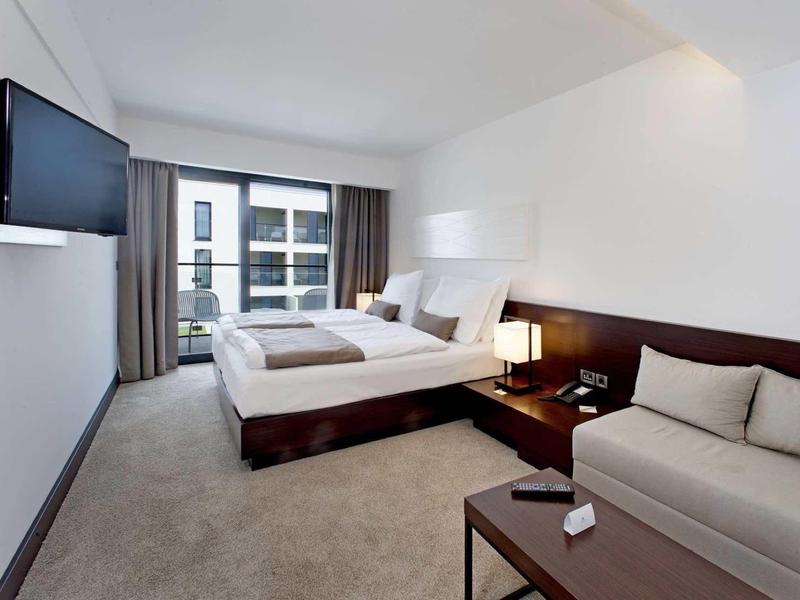 Jasny pokój hotelowy z podwójnym łóżkiem, sofą, telewizorem i balkonem z widokiem na budynek.