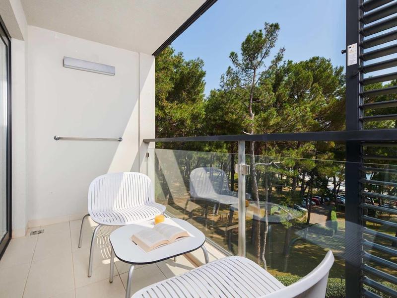 Balkon z białymi krzesłami i stołem, widok na zielone drzewa i ulicę.