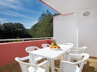 Balcón con mesa y sillas blancas, cesta de frutas, vista de árboles y cielo azul.