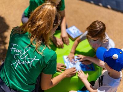 Animatrice en t-shirt vert joue à un jeu de cartes avec des enfants sur la plage.