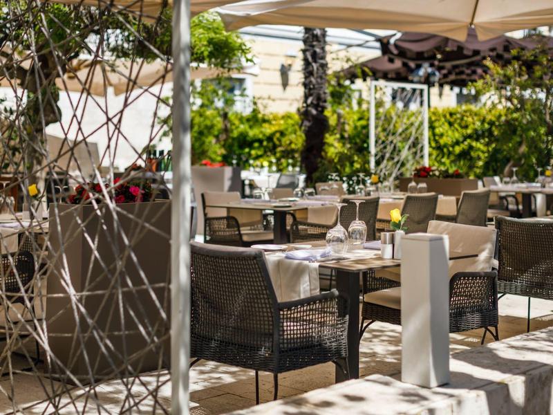 Venkovní restaurace s stolky a židlemi pod slunečními markýzami a bohatou zelení.