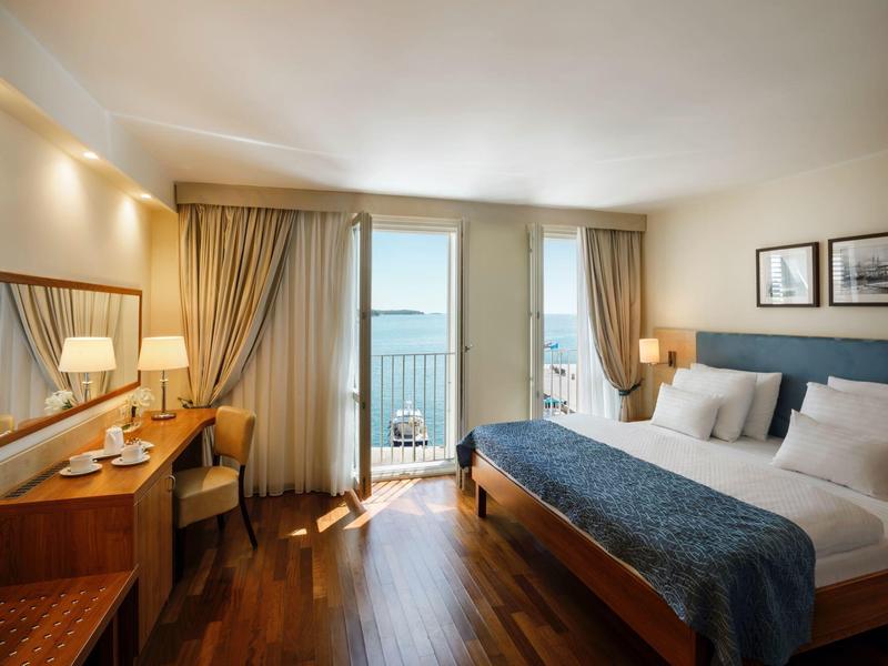 Světly hotelový pokoj s manželskou postelí, psacím stolem a balkonem s výhledem na moře.