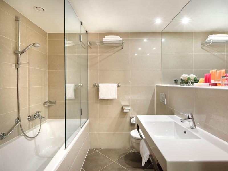 Moderní koupelna s vanou, skleněnou sprchovou stěnou, umyvadlem a ručníky.