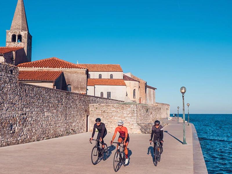 Tři cyklisté jedou po promenádě vedle kamenné zdi u moře pod jasnou modrou oblohou.