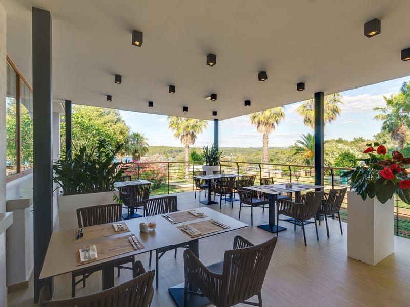 Terrazza coperta di un ristorante con tavoli e sedie circondata da verde.