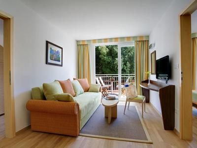 Světý obývací pokoj se zelenou pohovkou, televizí a přístupem na balkon s výhledem na zeleň.