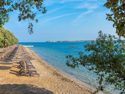 Leerer Sandstrand mit Sonnenliegen, blauem Meer und bewaldetem Ufer unter klarem Himmel.