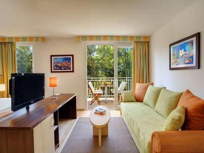 Moderní obývací pokoj se zelenou pohovkou, televizí a balkonem s výhledem na stromy.