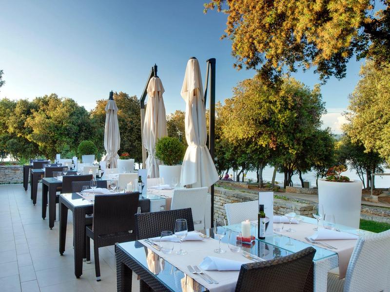 Venkovní část restaurace s prostřenými stoly a zavřenými slunečníky v zeleném prostředí.