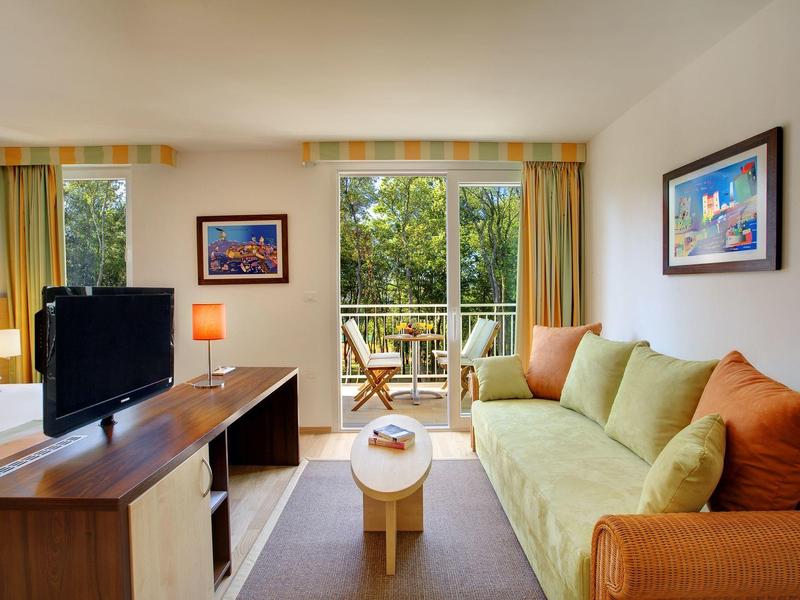 Moderní obývací pokoj se zelenou pohovkou, televizí a balkonem s výhledem na stromy.