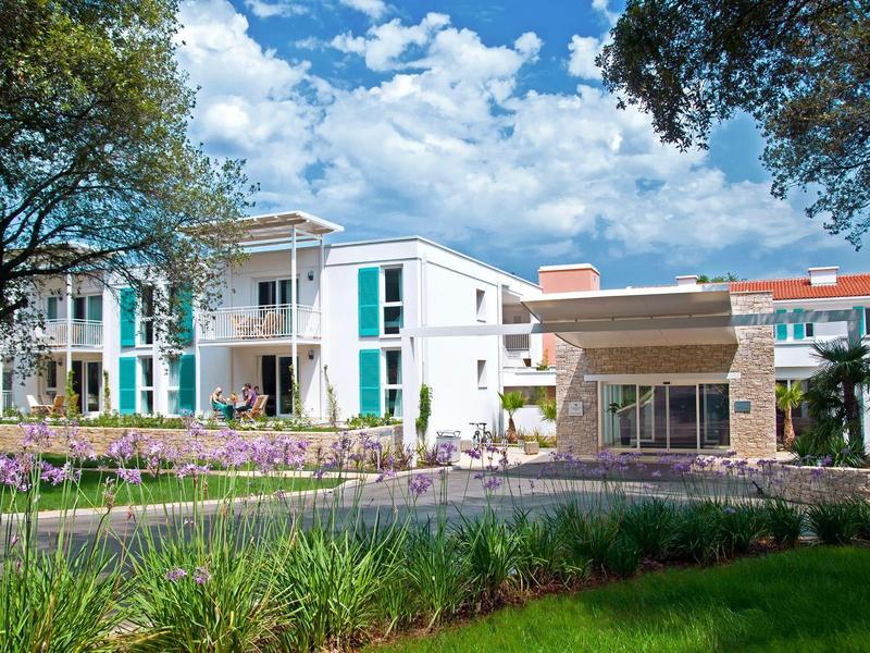 Moderní hotelová budova s bílými fasádami, velkými okny a okolní zelení pod modrou oblohou.