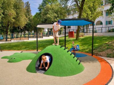 Enfant joue sur une structure de jeu crocodile verte avec des arbres et un bâtiment en arrière-plan.