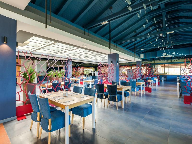 Restaurant lumineux avec chaises bleues et tables en bois, plafond haut avec structure bleue.