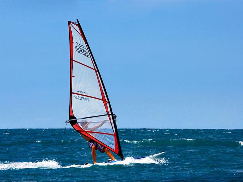 Un planchiste glisse sur la mer avec une voile rouge sous un ciel dégagé.