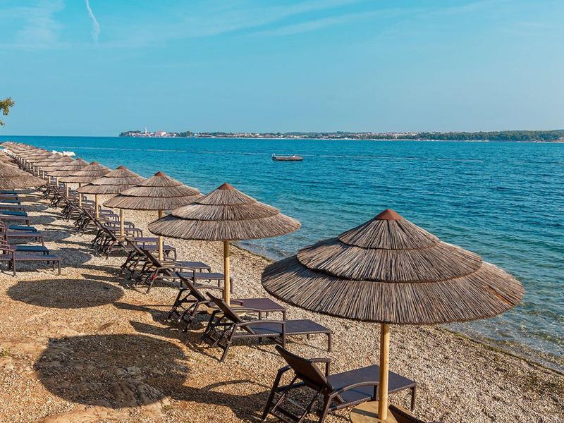 Rangée de transats avec parasols en paille sur une plage rocheuse au bord de la mer calme.