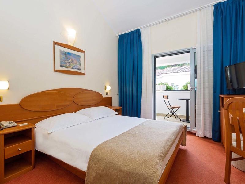 Chambre d'hôtel lumineuse avec lit double, table de chevet, bureau et balcon avec table et chaises.